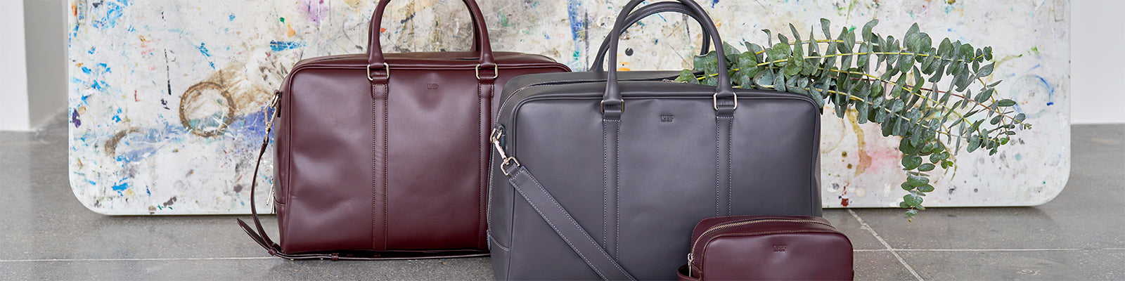 Two leather bags, one dark brown and one gray, are placed in front of a paint-splattered canvas. A smaller dark brown pouch and some greenery are also visible