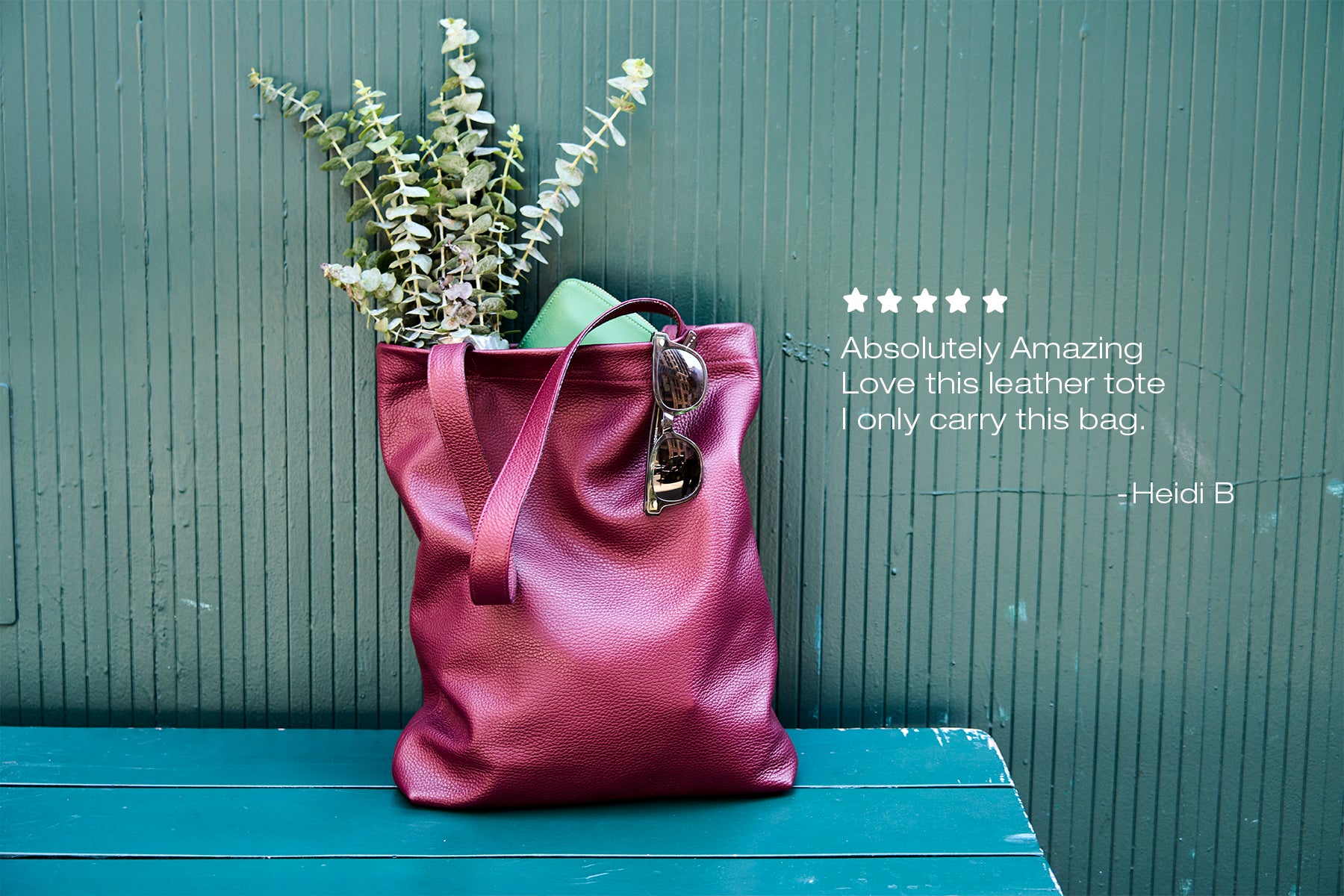 Deep burgundy leather tote with eucalyptus and sunglasses against a green wall.